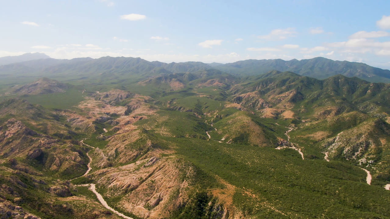 Second aerial frame in Cabo Pulmo, Baja California Sur, Mexico, with the scrub-covered foreground ridge more centered, the pale road tracing the valley floor, distant mountains, blue sky, and no visible people.