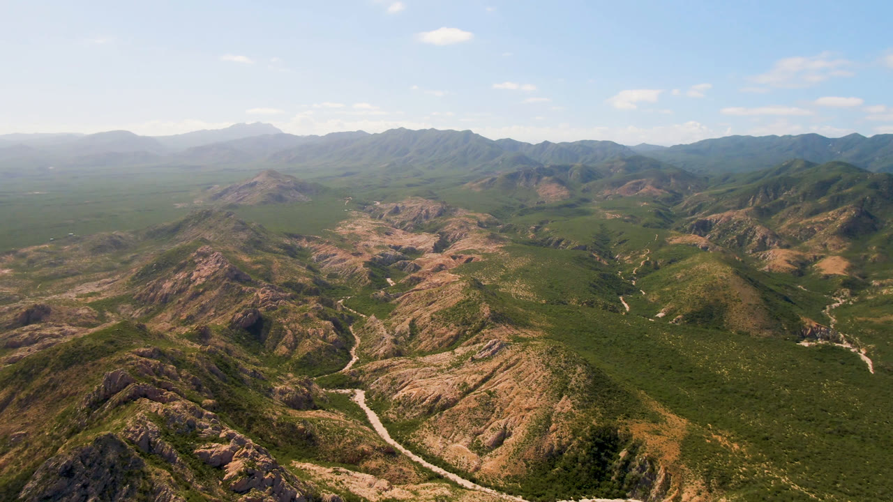 Early aerial frame over Cabo Pulmo mountain ridges, with a pale winding road or wash crossing the foreground valley, hazy peaks in the distance, and no visible people.
