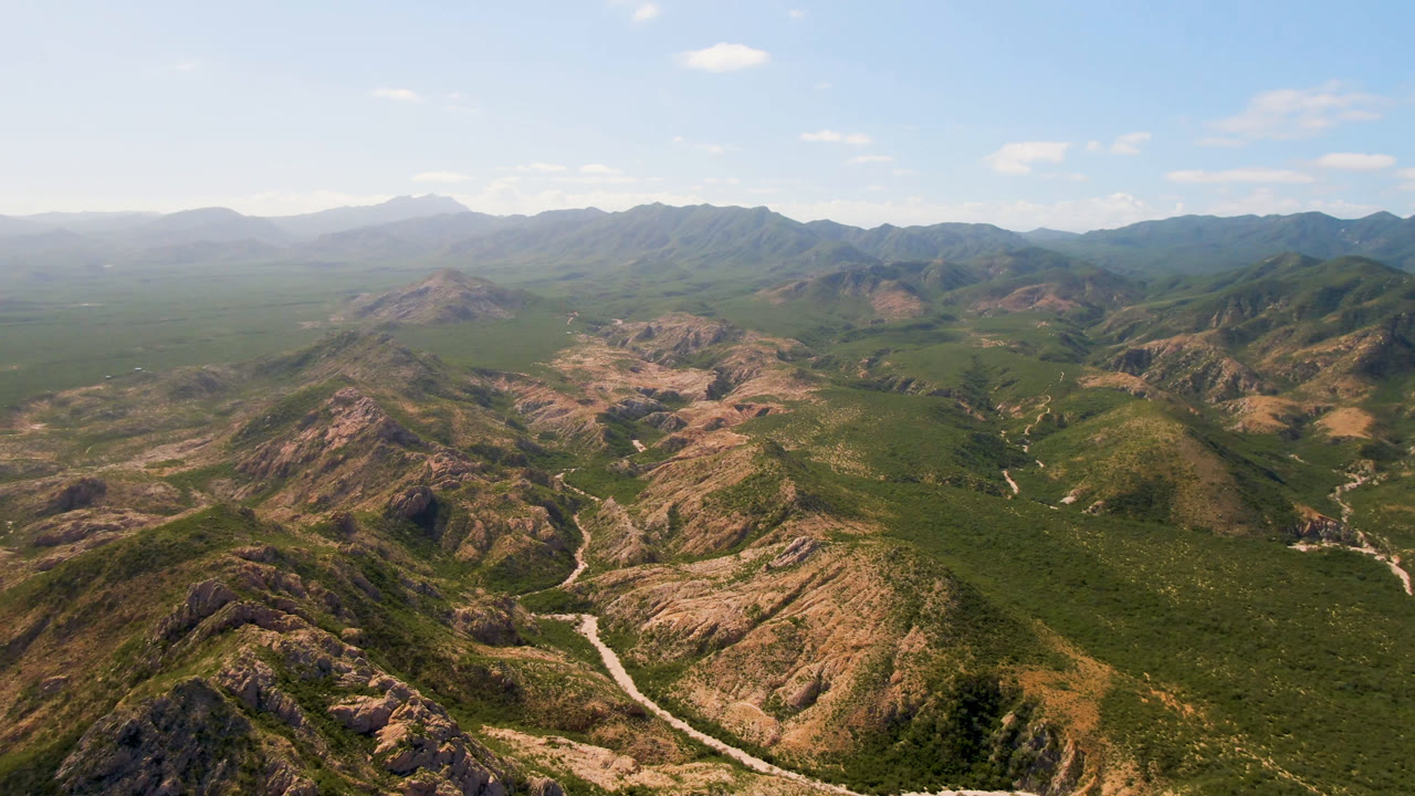 Aerial frame in Cabo Pulmo showing rugged green-brown hills, a pale winding track through the valley floor, layered background mountains, and no visible people.