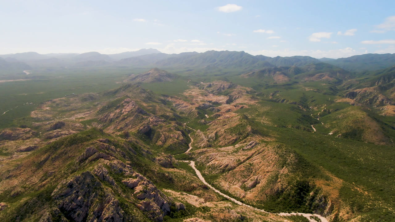 At 0:00, a wide aerial view over Cabo Pulmo shows rocky tan ridges, green desert vegetation, a pale winding road or wash below, distant mountains, and no visible people.