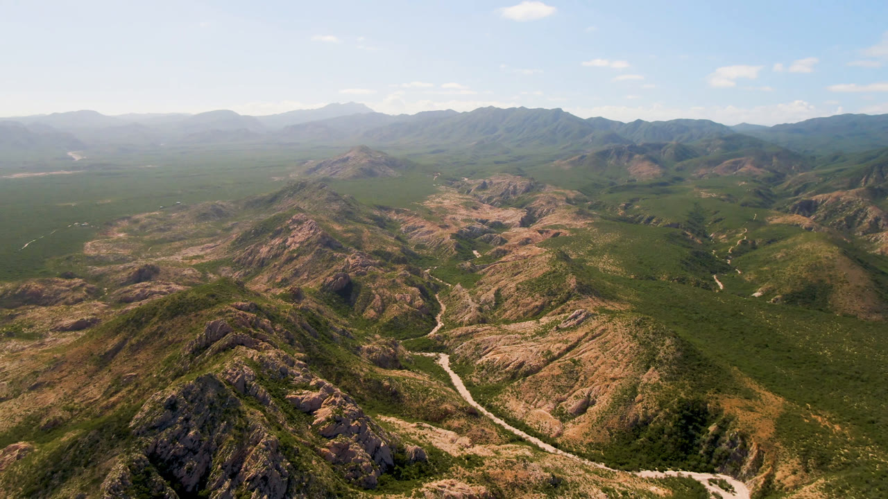 At 0:01, the Cabo Pulmo aerial view centers on layered rocky hills, broad green scrubland, a light-colored track in the valley, hazy background mountains, and no visible people.