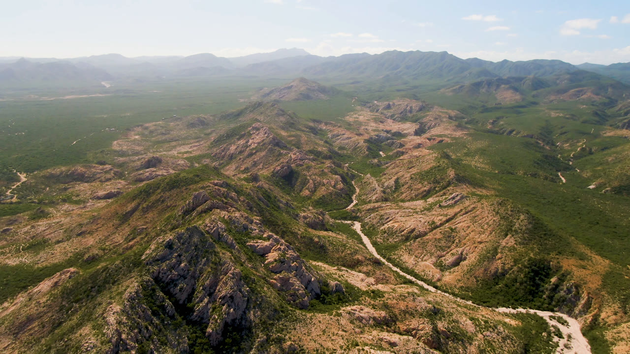 Aerial frame over Cabo Pulmo mountains with a rocky ridge in the foreground, a pale winding road in the valley, hazy distant ranges, and no visible people.