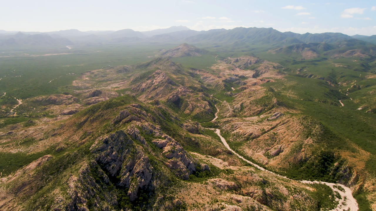 Aerial view in Cabo Pulmo with the central mountain ridge slightly shifted in frame, dry tan slopes, green vegetation, a pale valley route, and no visible people.