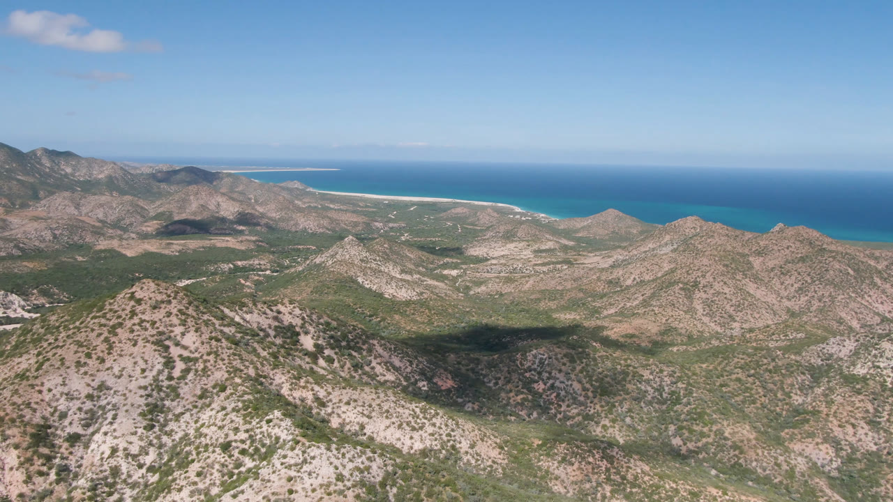 Opening frame over Cabo Pulmo, Baja California Sur, with scrub-covered rocky hills in front, a pale beach and blue ocean beyond, and no visible people.