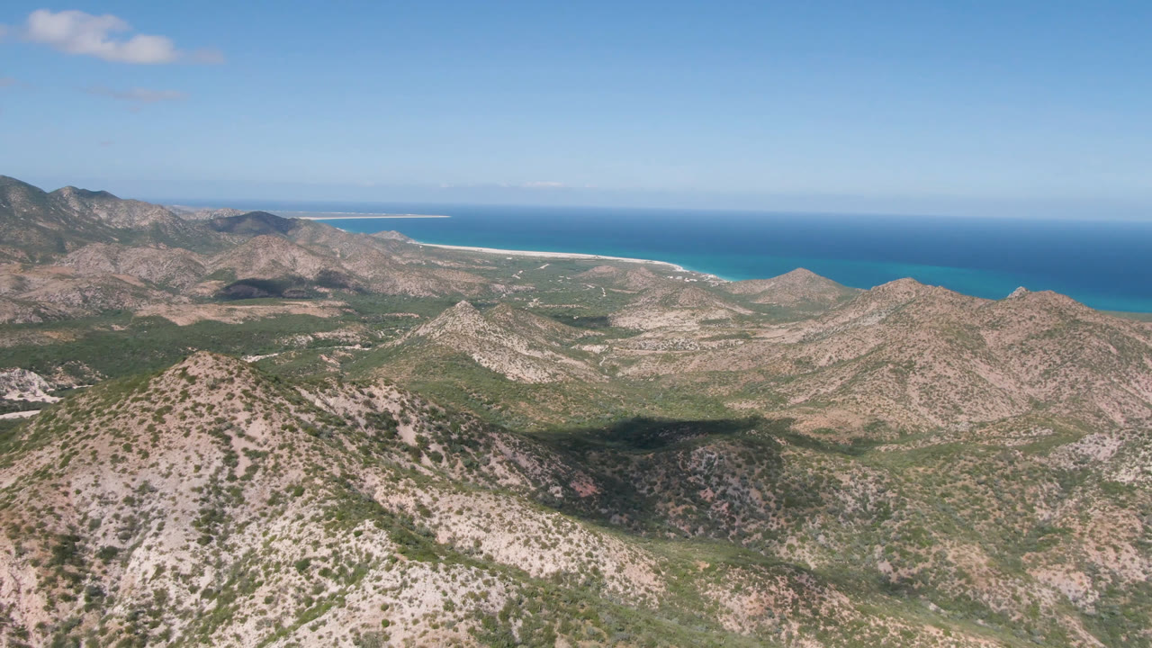 Second frame in Cabo Pulmo showing a central scrubby ridge, darker mountain slopes to the left, a turquoise shoreline, and no visible people.