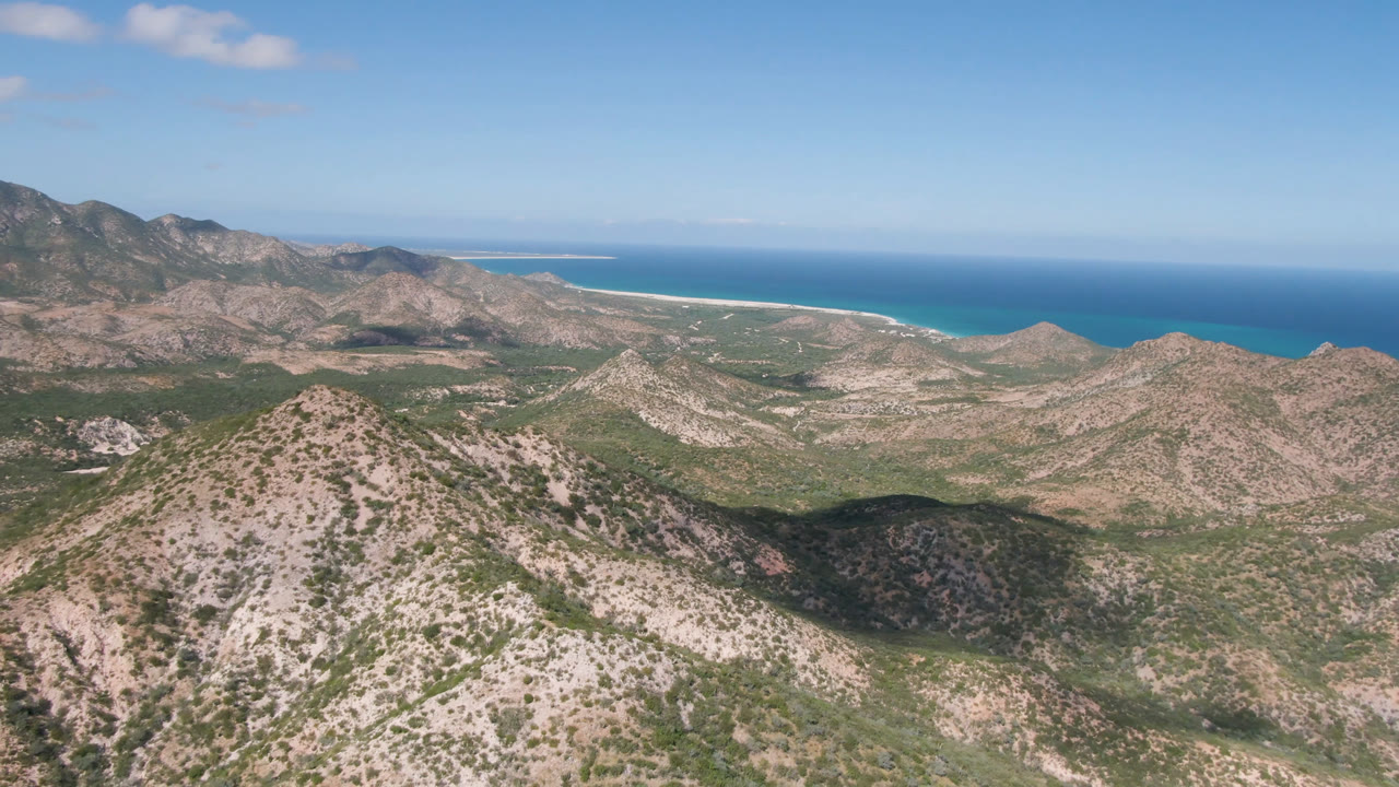 Wide aerial frame over Cabo Pulmo, Baja California Sur, with scrub-covered rocky hills in front, a turquoise ocean band beyond, and no visible people.