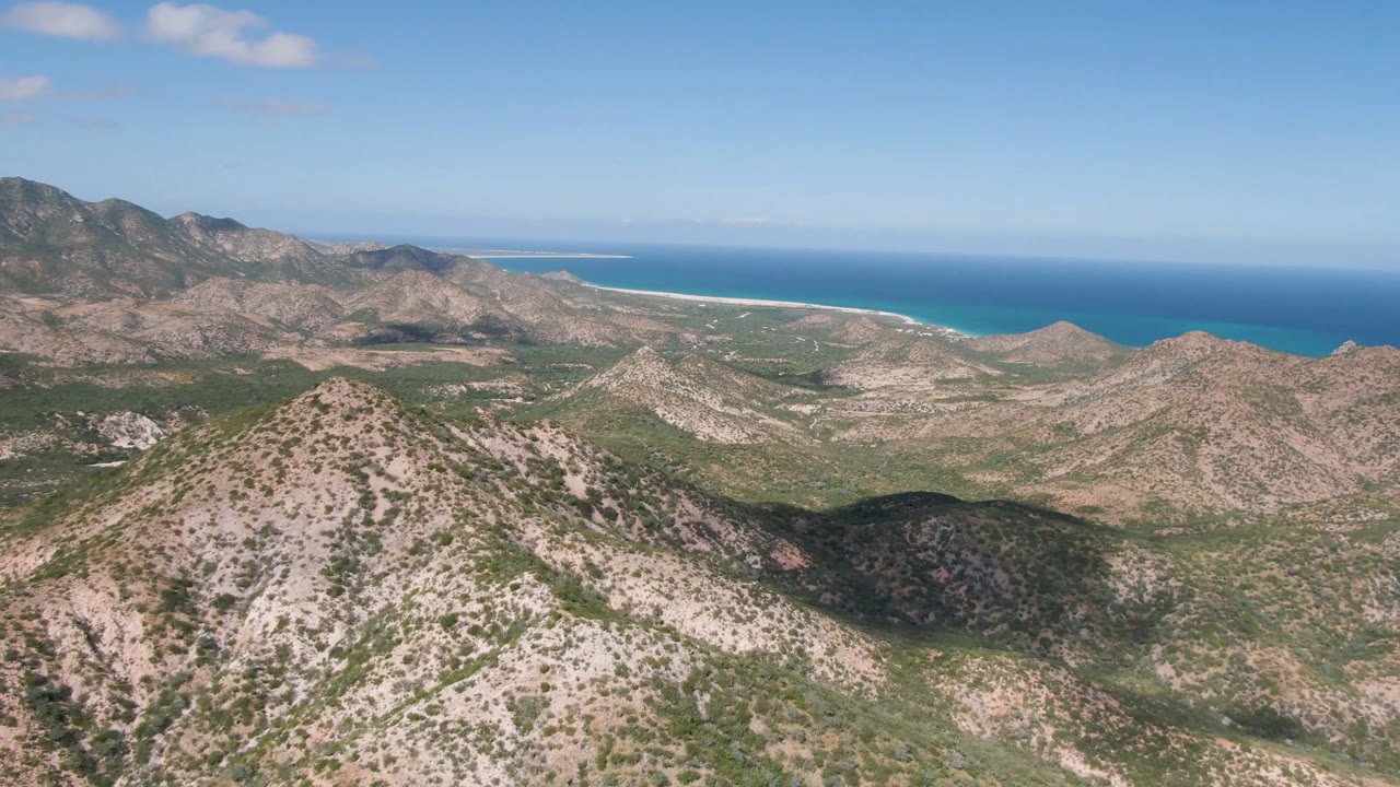 A high aerial view in Cabo Pulmo shows a central scrubby ridge, darker mountain slopes at left, blue ocean behind, and no visible people.