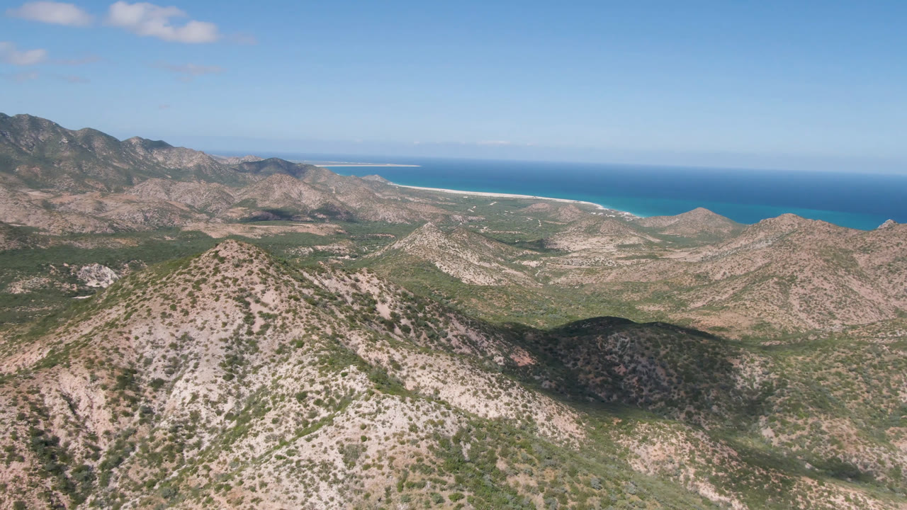 Broad aerial landscape with Baja hills, coastline, ocean, and scattered clouds.