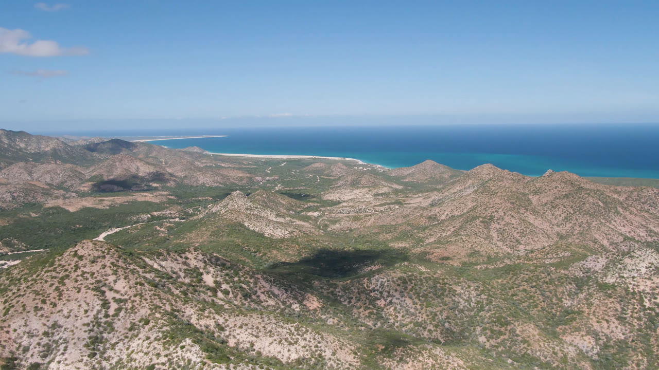 Opening aerial view over scrubby rocky hills toward a pale beach and blue ocean in Cabo Pulmo, Baja California Sur, Mexico, with no visible people.
