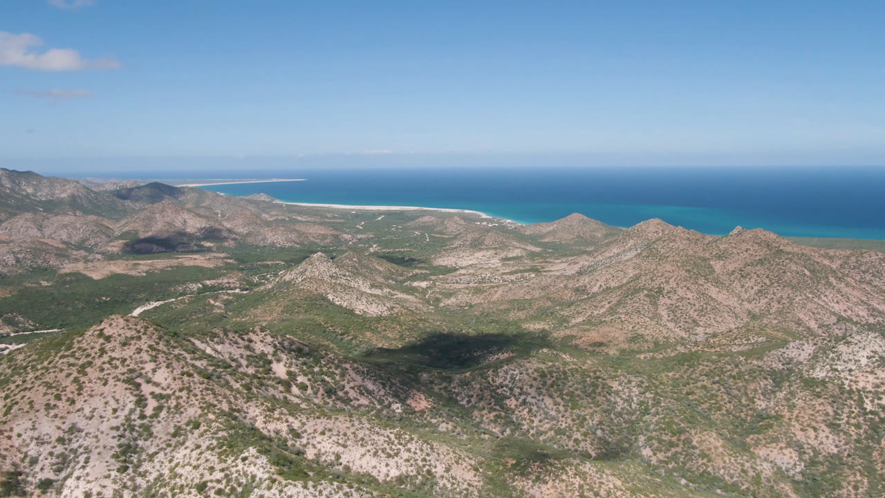 Wide aerial frame in Cabo Pulmo showing layered mountain ridges, sparse green vegetation, turquoise nearshore water, a thin sandy beach, and no visible people.