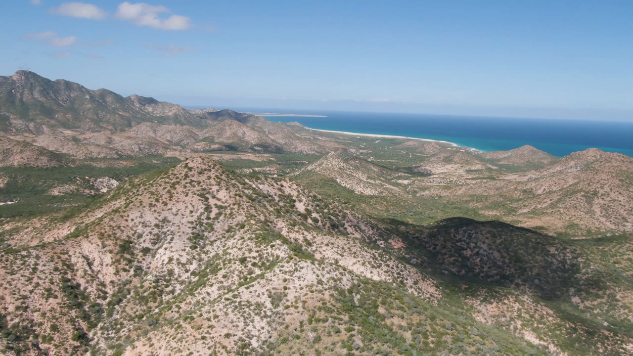 Aerial view of rocky desert mountains with ocean and beach beyond.