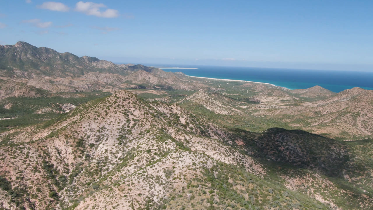 Aerial frame from Cabo Pulmo showing a rounded rocky ridge in front, dry green-brown terrain below, a pale beach strip, blue ocean, and no visible people.