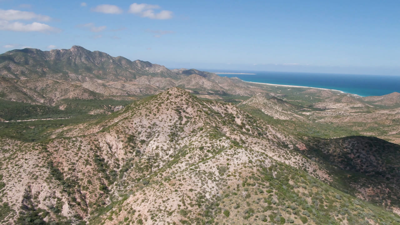 A wide Cabo Pulmo aerial frame shows the central ridge slightly closer in view, with mottled desert vegetation, distant mountains, a pale beach strip, blue ocean, and no visible people.