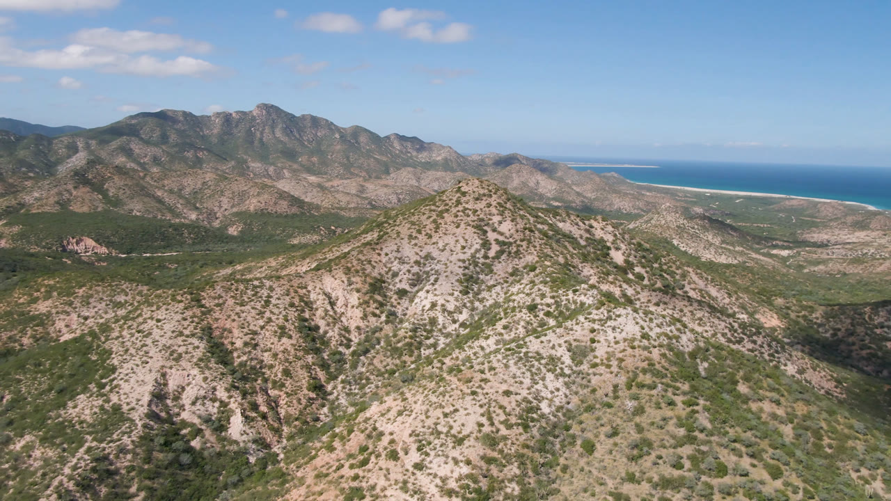 Aerial frame over a pale rocky ridge in Cabo Pulmo, Baja California Sur, with scrub-covered mountains, blue ocean on the right horizon, and no visible people.