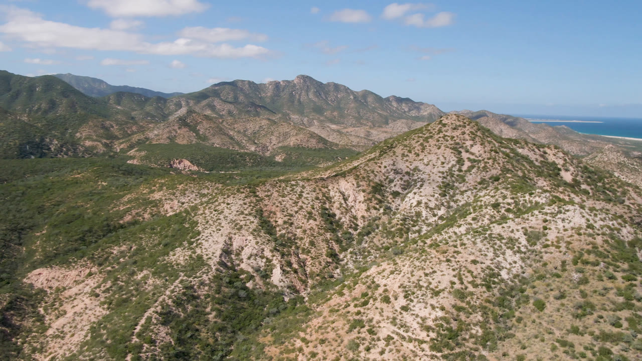 Aerial frame over Cabo Pulmo, Baja California Sur showing a pale shrub-covered ridge, darker green valley vegetation, distant rugged mountains, blue sky, and no visible people.