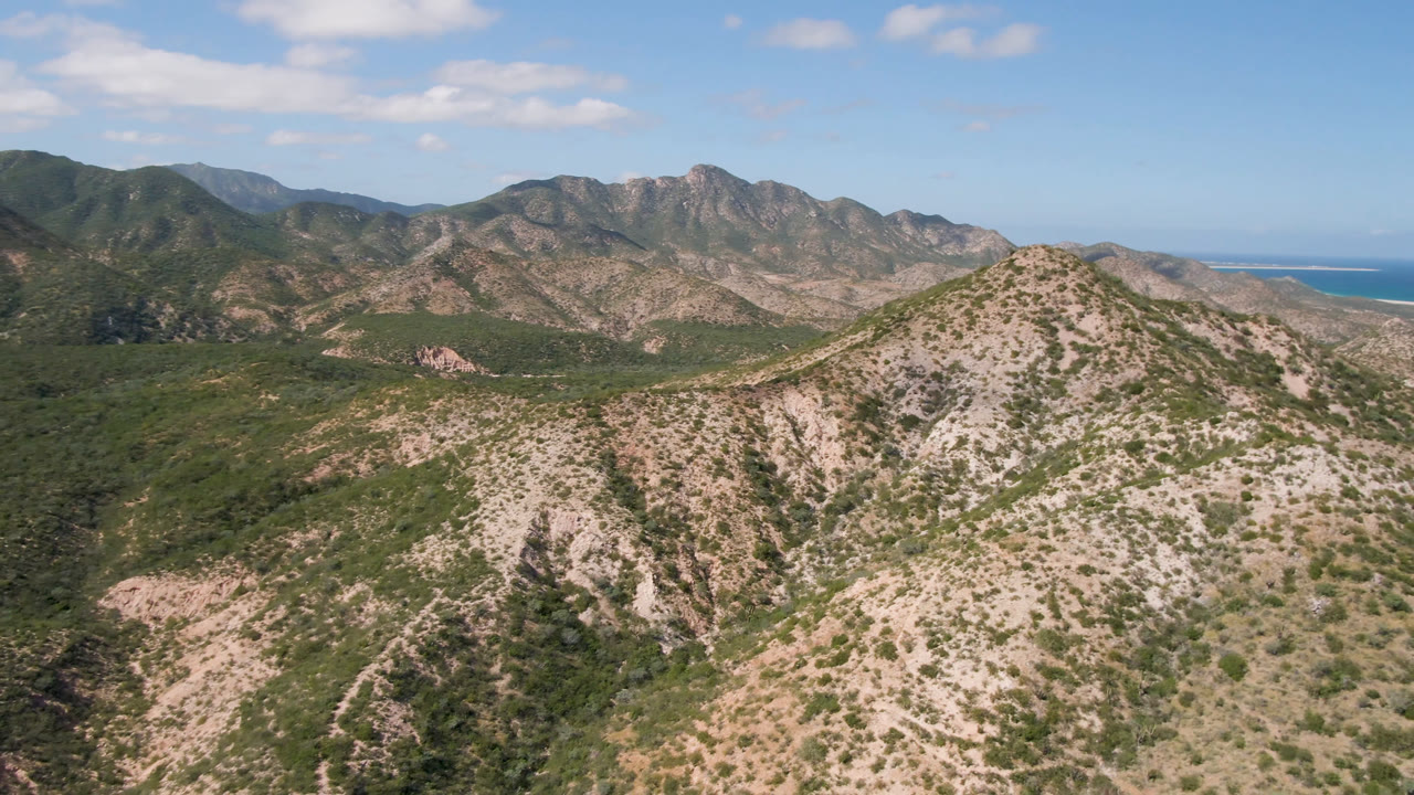 Aerial view in Cabo Pulmo with the foreground ridge centered slightly lower, a broad green valley below the mountain range, small clouds overhead, and no visible people.