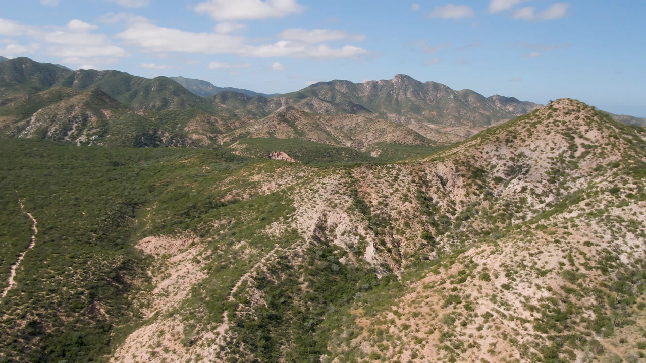 Opening aerial frame over Cabo Pulmo desert mountains, with green scrub on rocky slopes, distant ridges, scattered clouds, and no visible people.