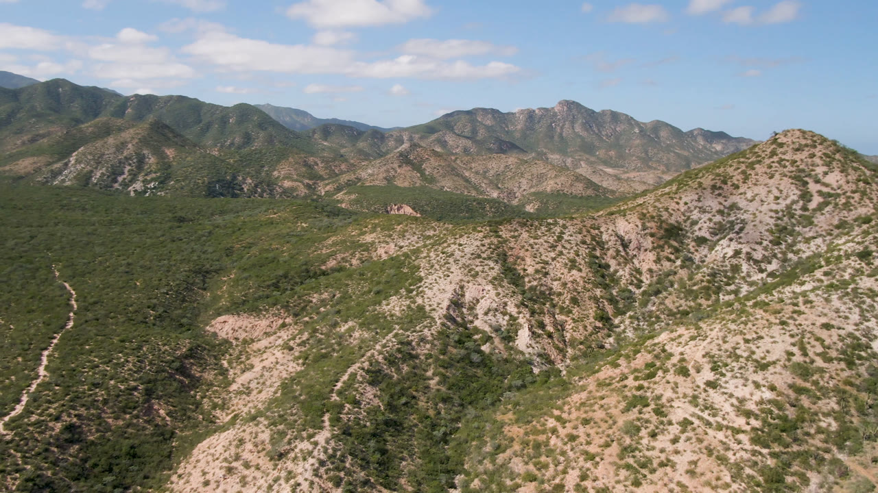 Aerial frame in Cabo Pulmo showing a pale rocky ridge in the foreground, dense green scrub in the valley, distant mountains, and no visible people.