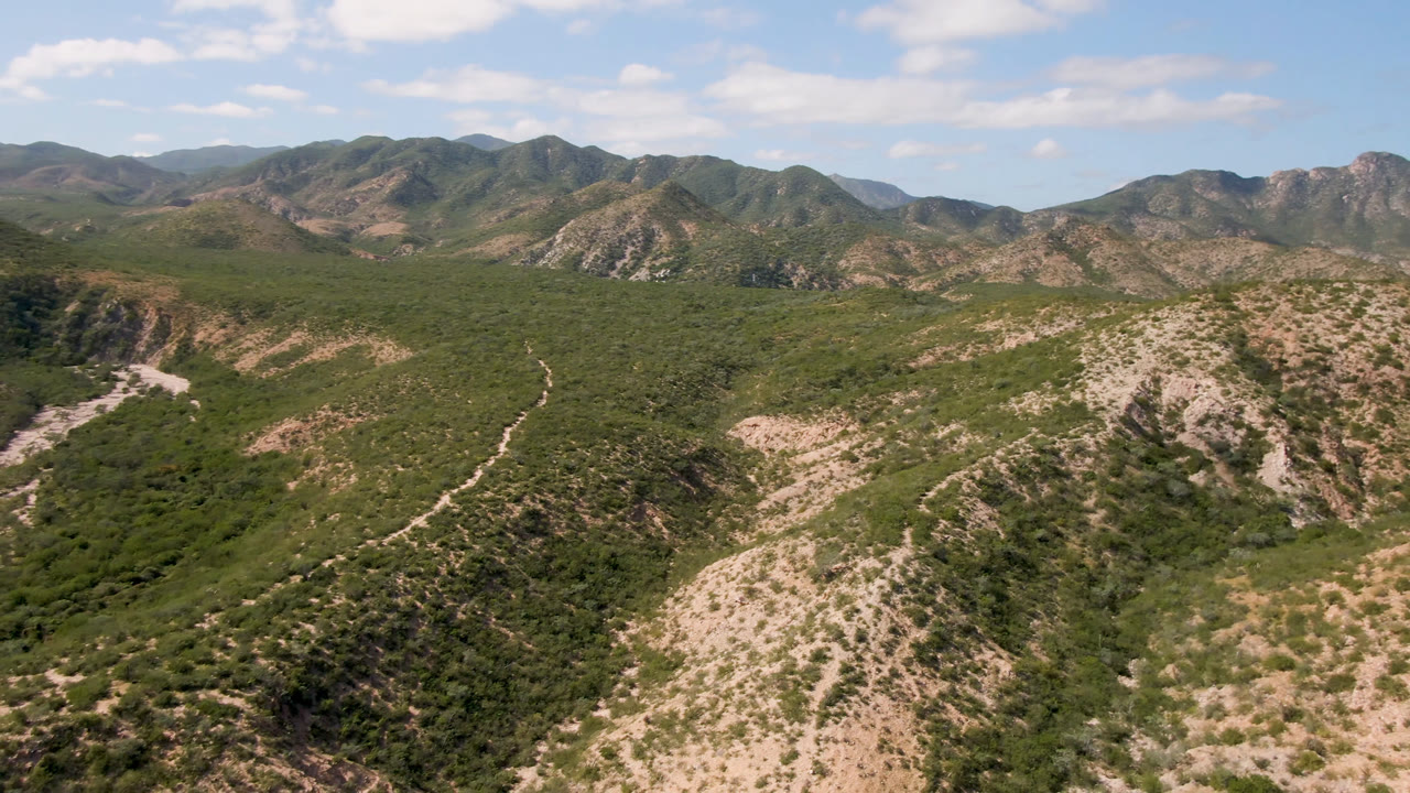 Aerial view in Cabo Pulmo showing a scrubby valley between rocky hills, with distant ridgelines under scattered clouds and no visible people.