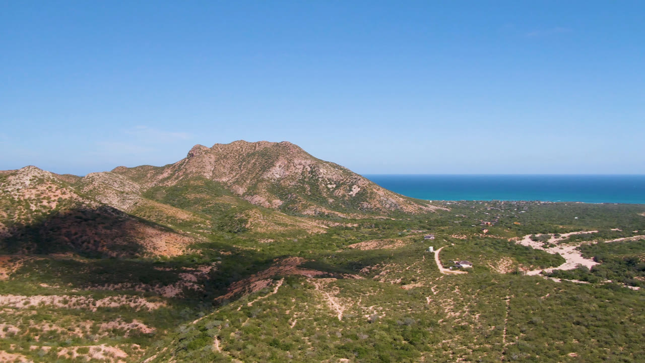 Cabo Pulmo aerial frame with the central rocky ridge, green-brown scrubland, pale dirt roads, and the ocean band visible beyond the mountains, with no visible people.