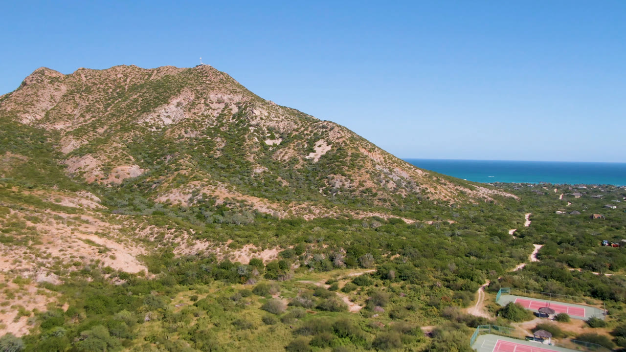 Opening aerial frame over Cabo Pulmo, Baja California Sur, showing a tan and green mountain ridge, scrubland, dirt tracks, a small sports court near the lower edge, blue ocean, clear sky, and no visible people.