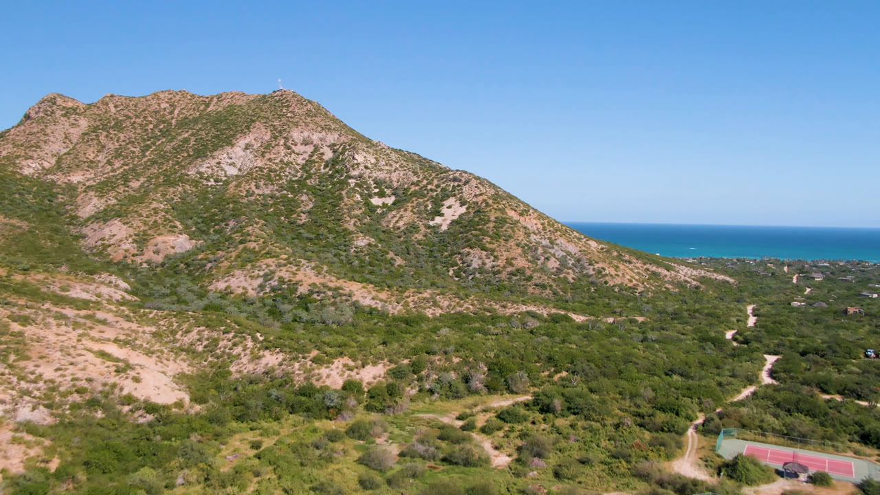 Aerial frame in Cabo Pulmo with the rocky mountain centered higher in view, green desert scrub below, a pale winding road at right, blue ocean behind the hills, clear sky, and no visible people.