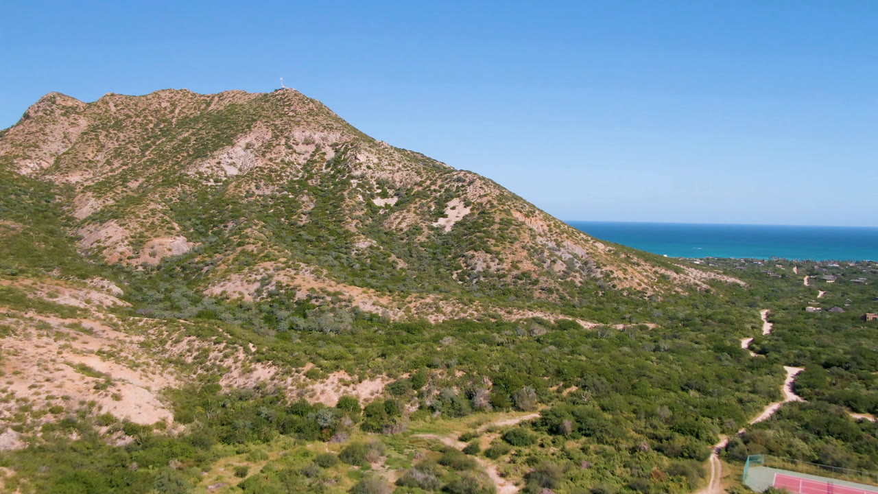 Aerial landscape with mountain slopes, scrubland roads, ocean, and clear blue sky.