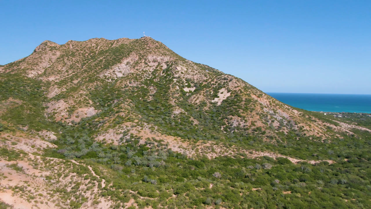 A wide aerial frame from Cabo Pulmo shows scrub-covered ridges, pale exposed rock, a small dirt route across the lower slope, blue ocean beyond, and no visible people.