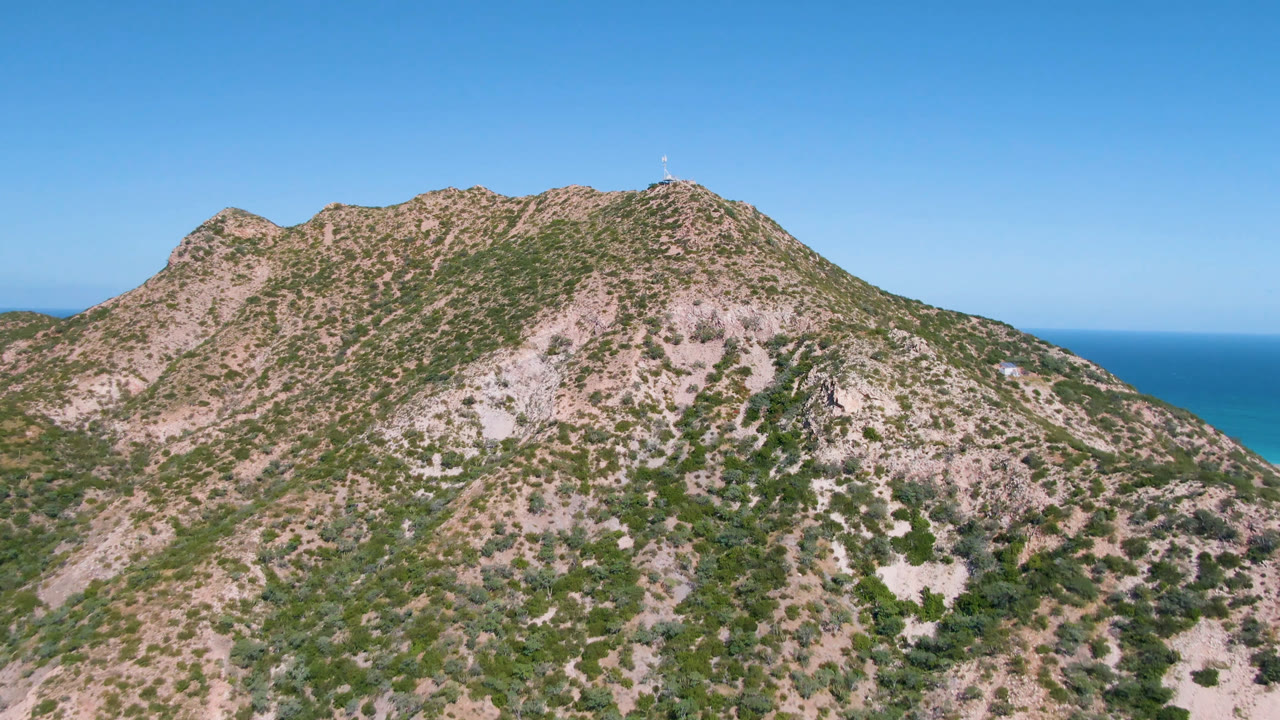 Mountain crest with scrub vegetation and bright blue ocean under clear sky.