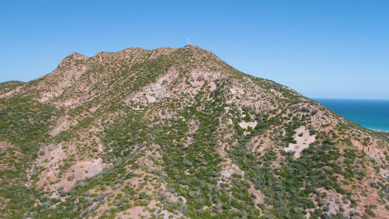 Opening frame of a scrub-covered rocky mountain in Cabo Pulmo, Baja California Sur, with a blue ocean strip and clear sky behind it, no visible people.