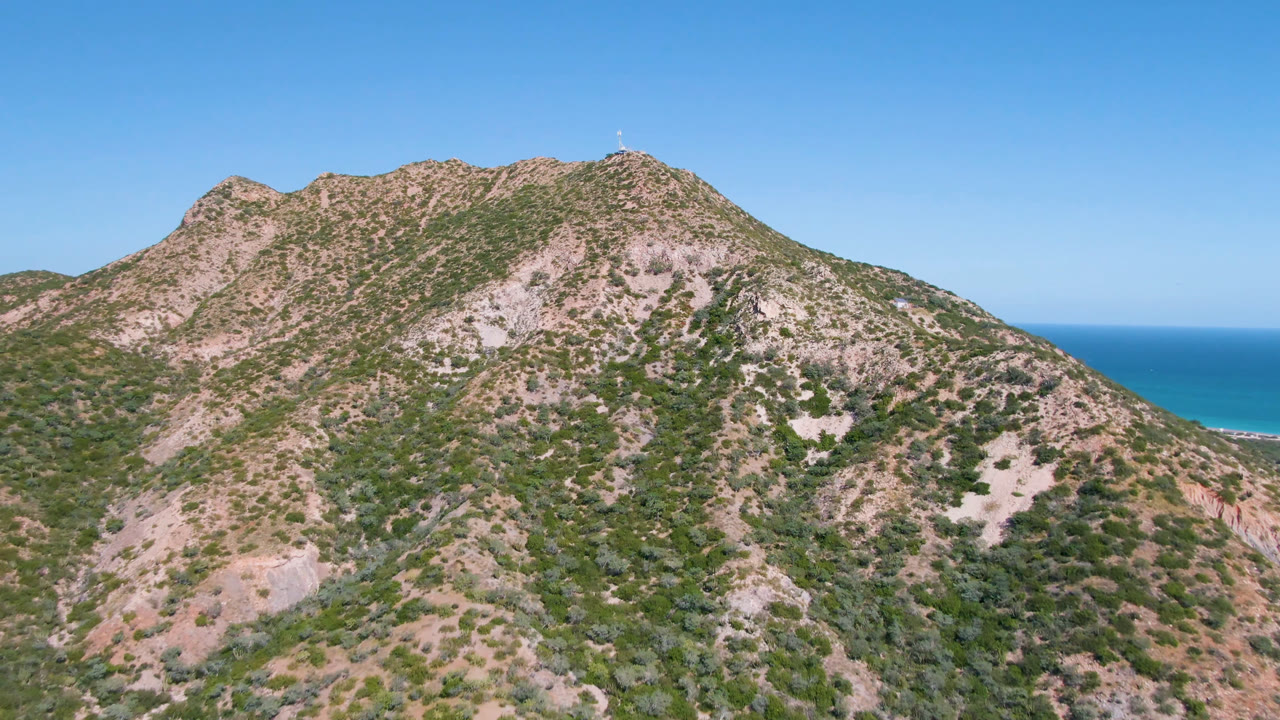 Early aerial frame in Cabo Pulmo showing the rugged mountain ridge centered in frame, with green scrub, pale rock patches, blue ocean, and no visible people.