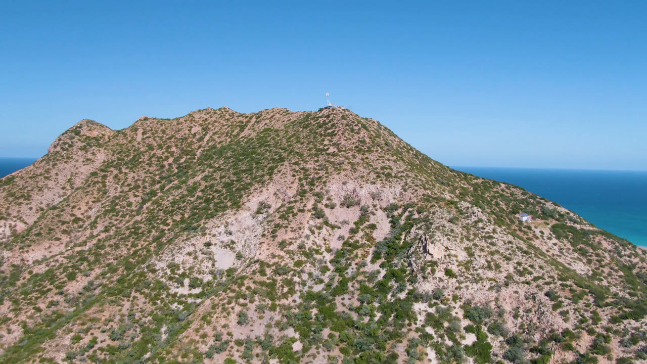 Aerial frame from Cabo Pulmo centers the rugged mountain ridge, with patchy green vegetation, pale rock faces, ocean beyond the saddle, and no visible people.