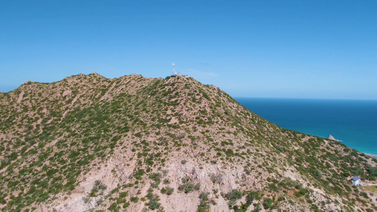 A Cabo Pulmo aerial frame shows the rocky ridge more centered, with sparse green scrub, blue water behind it, and a small white structure near the top, with no visible people.