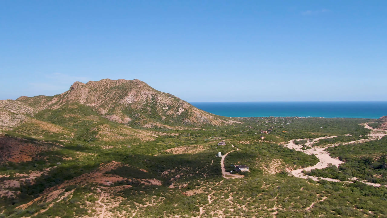 Opening aerial frame over Cabo Pulmo, Baja California Sur, showing rocky green-brown mountains, pale dirt tracks, blue ocean beyond, and no visible people.