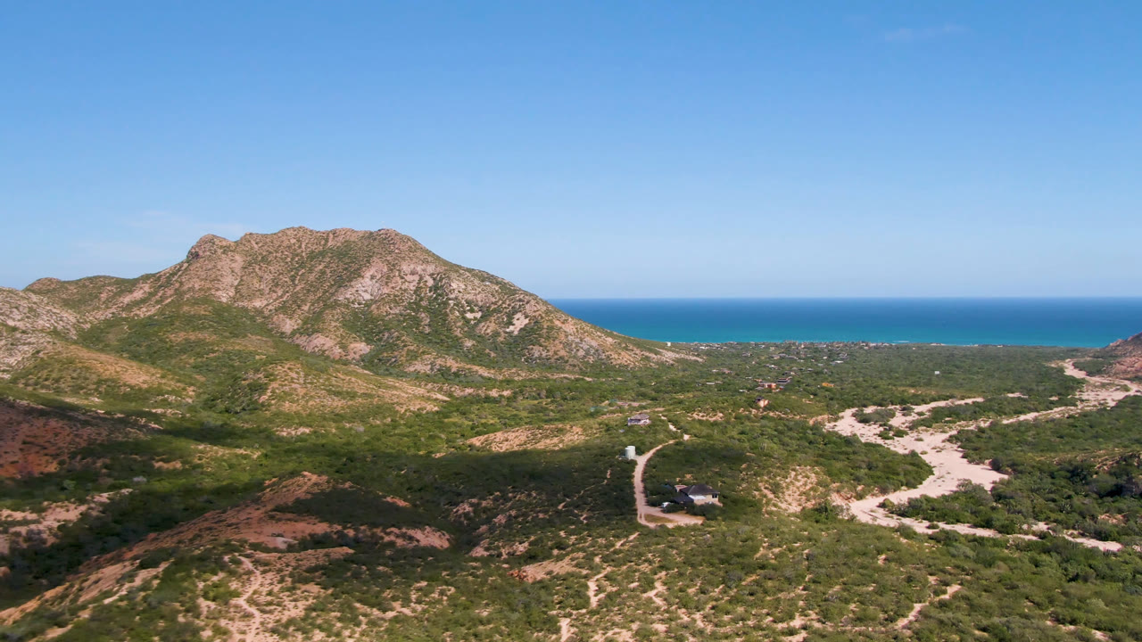 Second aerial frame in Cabo Pulmo with the mountain ridge centered, a winding pale road below, scattered desert vegetation, ocean horizon, and no visible people.