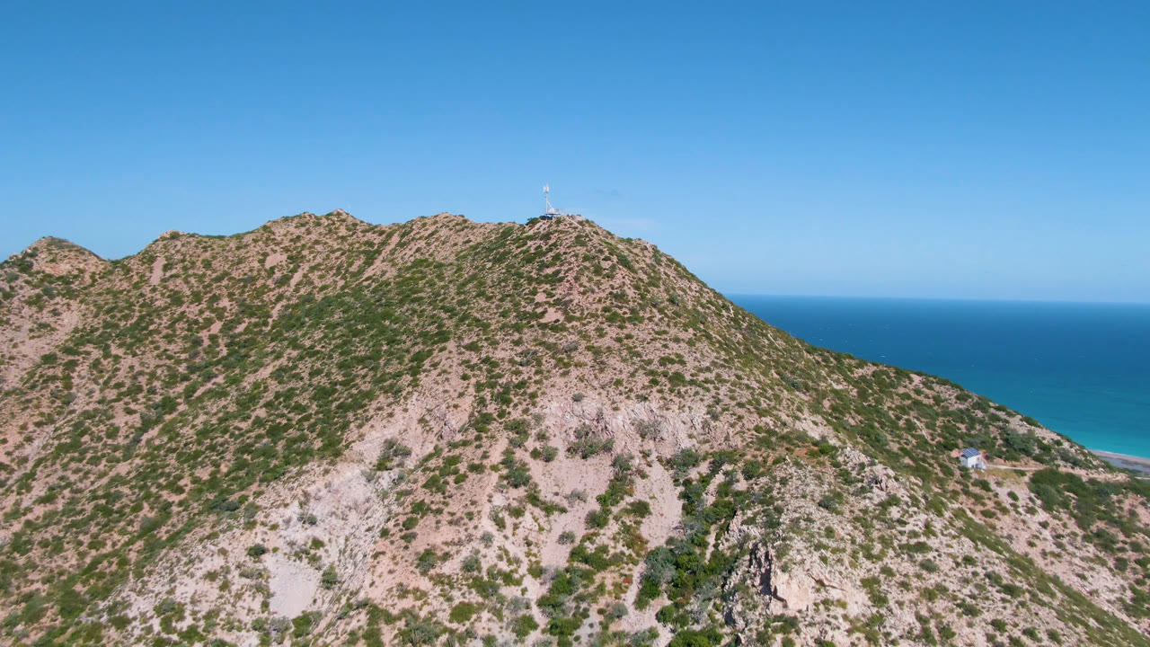 Second frame holds on the Cabo Pulmo ridge, with the cross near the skyline, pale rock faces below, deep blue ocean in back, and no visible people.
