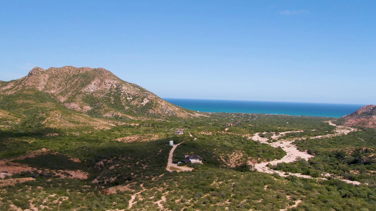 Aerial frame in Cabo Pulmo with the mountain centered over scrub-covered terrain, a pale road curving through the lower frame, the ocean behind, and no visible people.