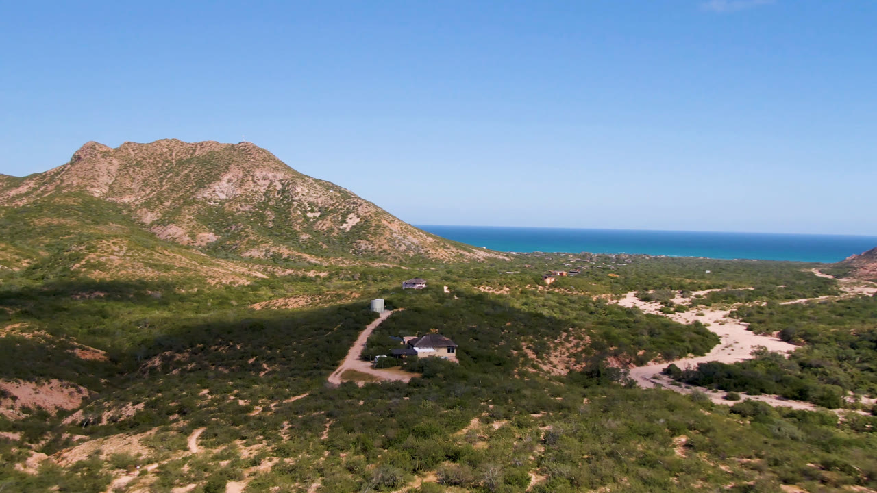 Aerial frame in Cabo Pulmo, Baja California Sur with the rocky hill centered, dark green scrub in the foreground, a small distant structure, blue ocean, clear sky, and no visible people.