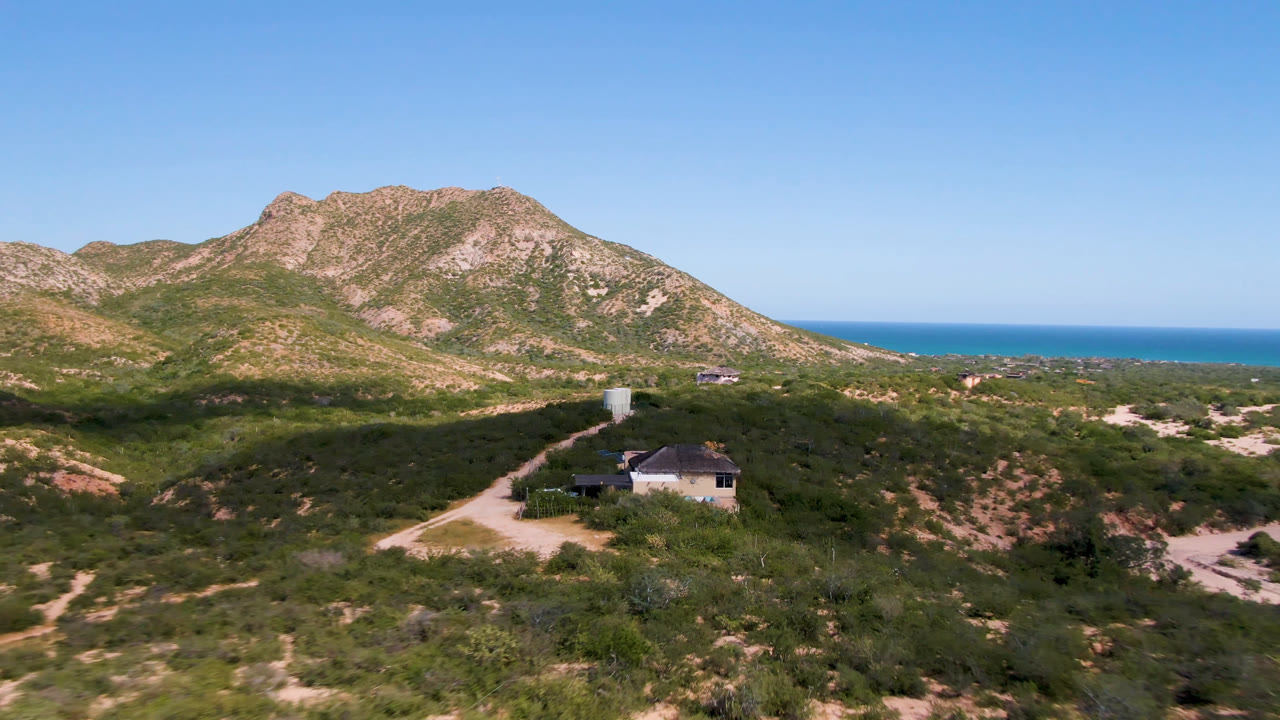 Opening aerial frame in Cabo Pulmo, Baja California Sur, showing a small house beside a dirt road below rocky hills with the ocean horizon beyond and no visible people.
