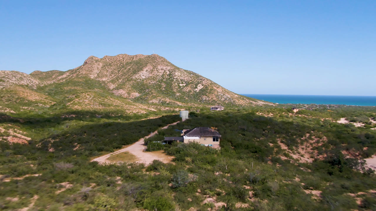 At one second, the Cabo Pulmo drone view keeps the house low in frame while the dirt track, scrub-covered slope, rocky ridge, and ocean horizon remain visible with no visible people.