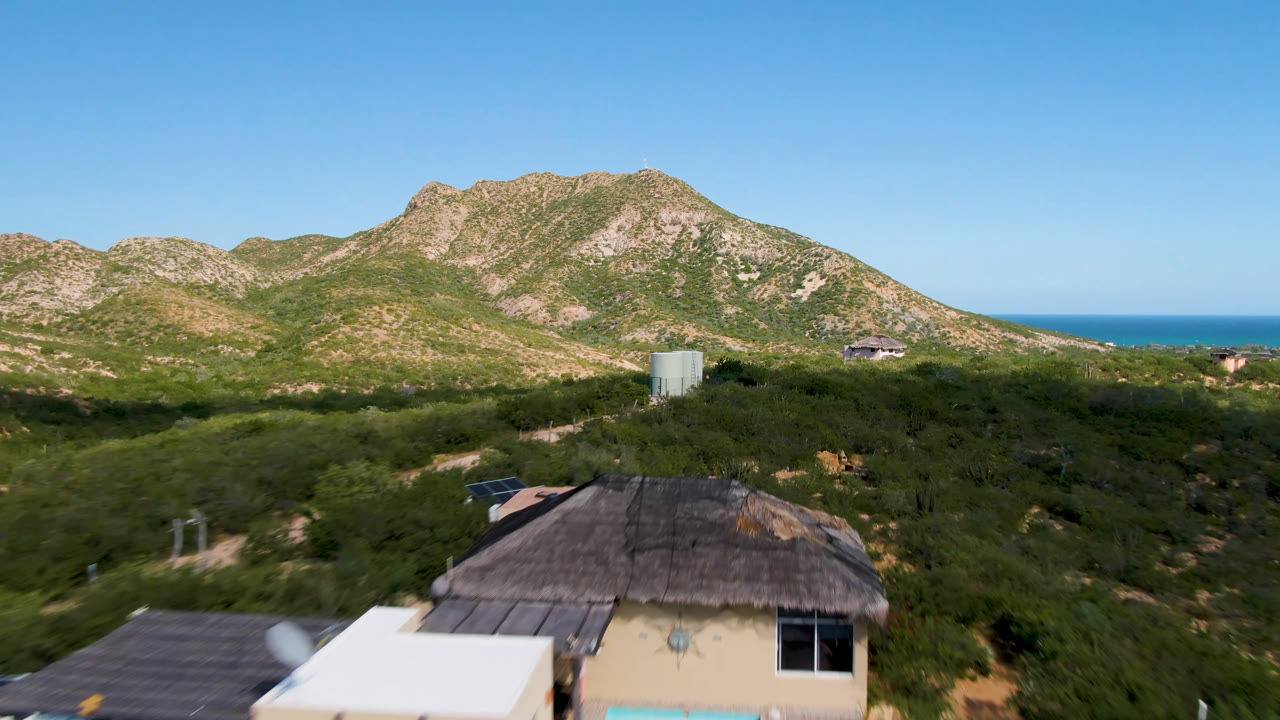 At 0:00, an aerial frame in Cabo Pulmo looks past a thatched roof and white rooftop area toward scrubland, a pale water tank, rocky hills, blue ocean, and no visible people.