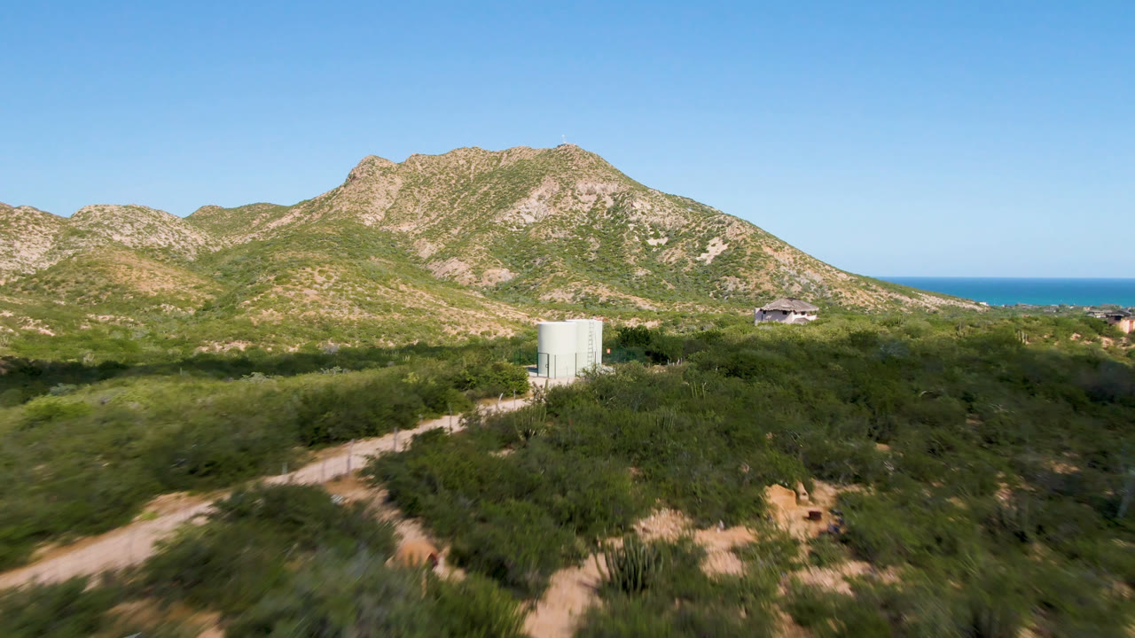 Water tank in a brushy field with dirt paths, mountains, ocean, and blue sky.