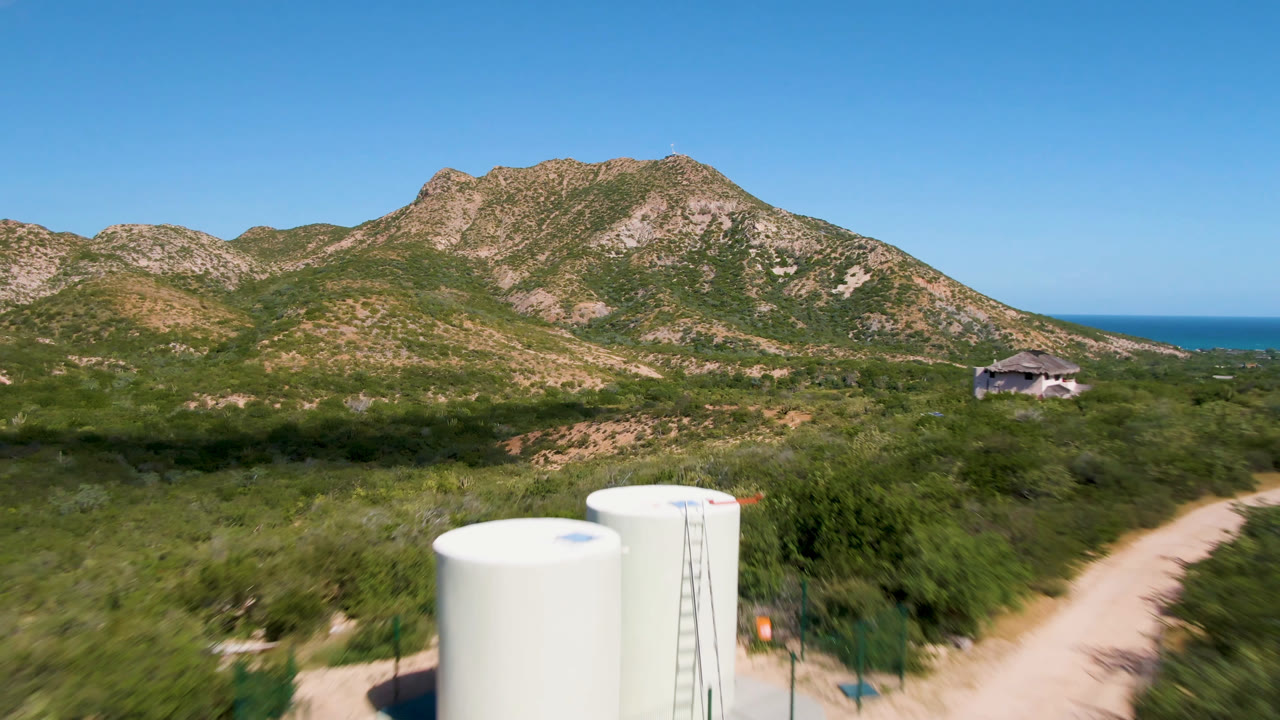 White cylindrical water tanks sit in the lower foreground of a Cabo Pulmo aerial view with scrubland, a dirt road, rocky mountains, blue ocean on the horizon, and no visible people.