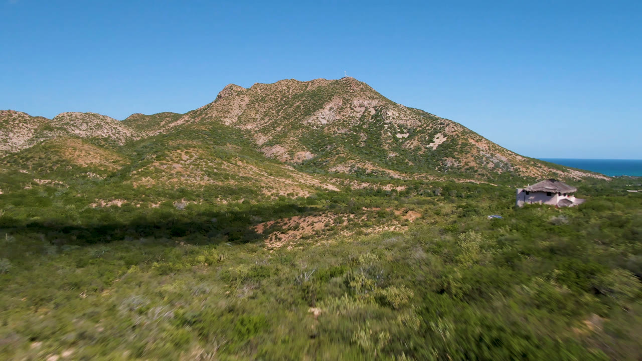 A Cabo Pulmo aerial frame looks across a dirt road and dense green scrub toward rocky mountains, a thin blue ocean horizon, a small structure at right, and no visible people.