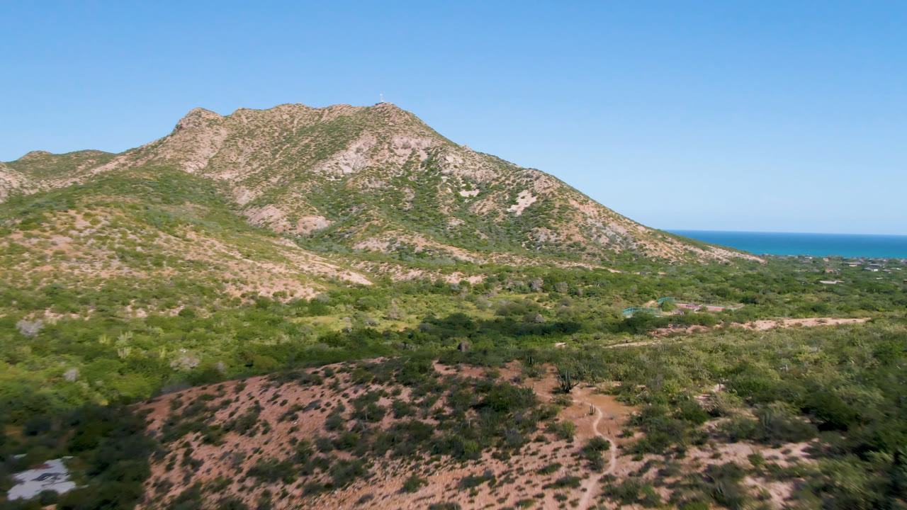 Second frame in Cabo Pulmo keeps the rocky mountain centered, with scrubland and dirt paths below, blue water at the horizon, and no visible people.