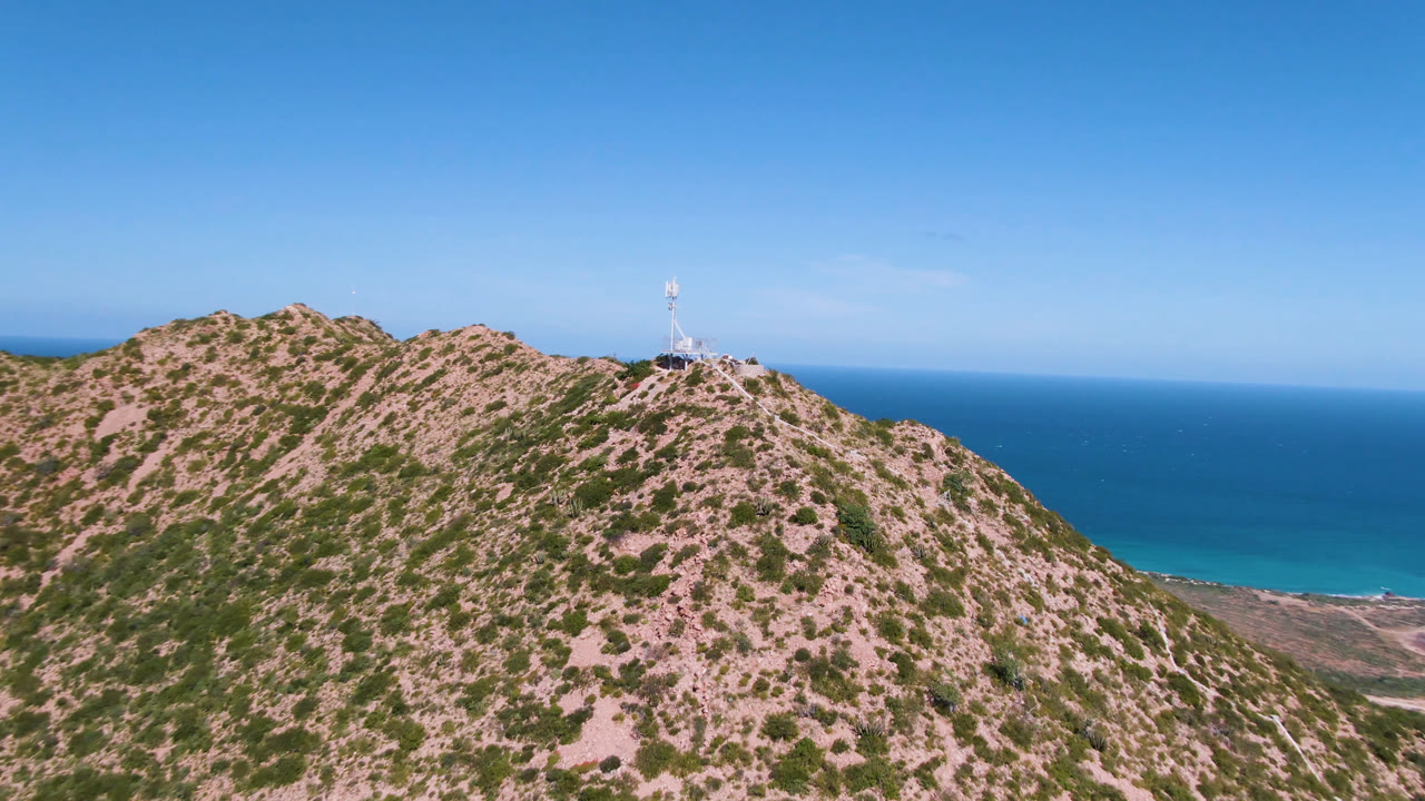 Aerial frame from Cabo Pulmo, Baja California Sur, with the rocky ridge centered, the lighthouse structure near the upper crest, turquoise shoreline water at right, and no visible people.