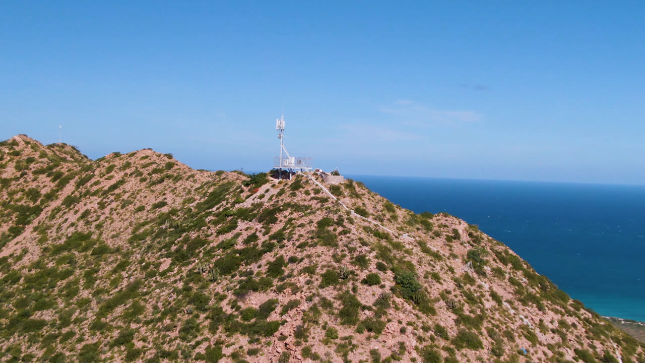 Wide aerial frame of a rocky scrub-covered hill in Cabo Pulmo, Baja California Sur, with a small tower near the ridgeline, blue ocean beyond, clear sky, and no visible people.