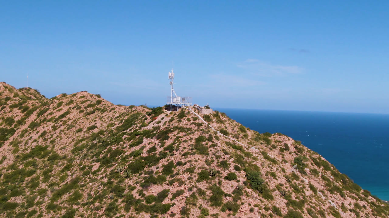 Aerial view of a tan Cabo Pulmo ridge with patchy green vegetation, a tower on the upper slope, blue water behind the hill, and no visible people.