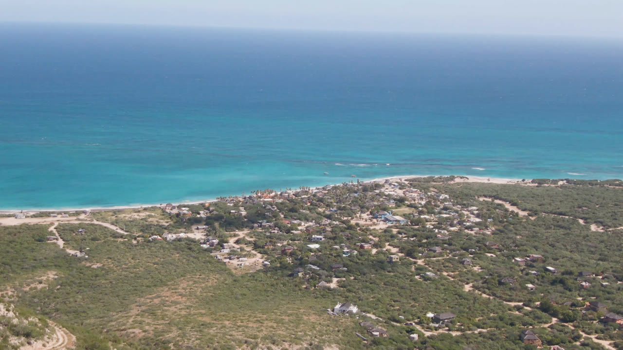 Aerial view over Cabo Pulmo, Baja California Sur, with turquoise ocean beyond a sandy beach, scrubby hillside roads, scattered buildings, and no visible people.
