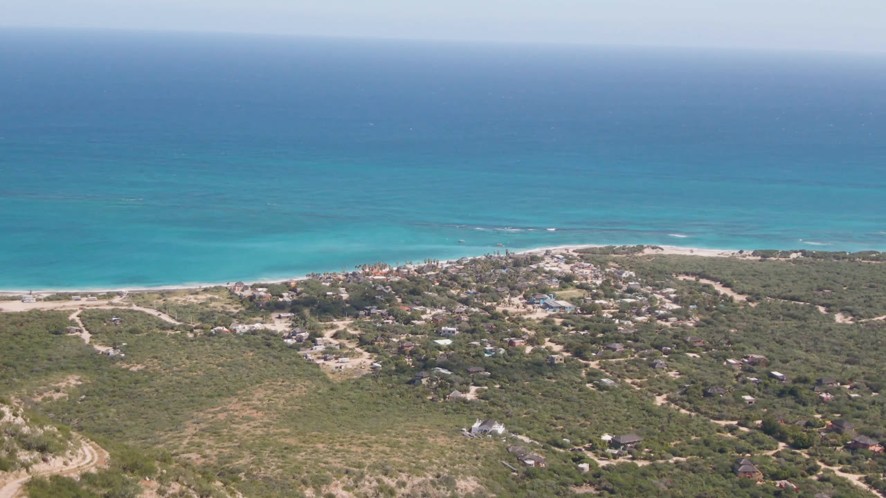 High aerial frame of Cabo Pulmo with the ocean horizon high in the shot, pale surf at the beach, dirt tracks through green-brown scrub, and no visible people.